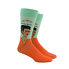 Shown on a leg form, a pair of Hot Sox cotton men’s crew socks with the artist Frida Kahlo in an orange shirt with a soft green background and her name in text above her