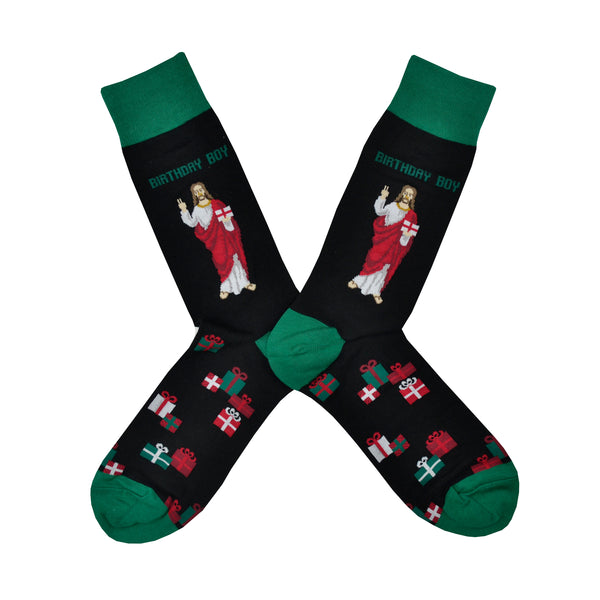 Shown in a flatlay, a pair of men's cotton and polyester HotSox brand black crew socks with a green heel, toe, and cuff. The leg of the sock features a cartoon design of Jesus with the words 'Birthday Boy' above. The foot of the sock features little holiday wrapped gifts.