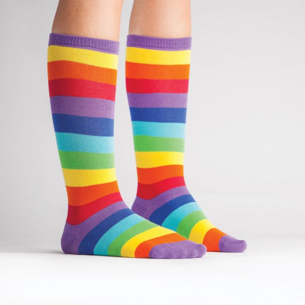 Shown on a light skinned child model, a pair of children's knee high Sock it to Me brand cotton socks with a purple heel, toe, and cuff. The rest of the sock is a classic rainbow stripe in yellow, orange, red, purple, indigo, blue, and green.
