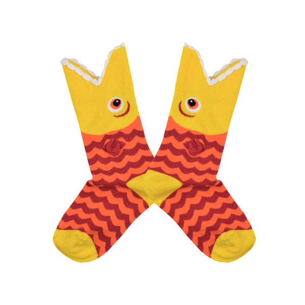 Shown in a flatlay, a pair of K.Bell brand kids cotton crew socks with a yellow heel, toe, and cuff and an orange and red sock. The top of the sock features a mouth shaped top with a cartoon fish eye and the body of the sock is red and orange zig zag.