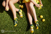 Two people sit on grass peeling lemons while wearing a mismatched pair of Many Morning's unisex cotton ankle socks that show lemons on sock and a big lemon peel on the other sock. 