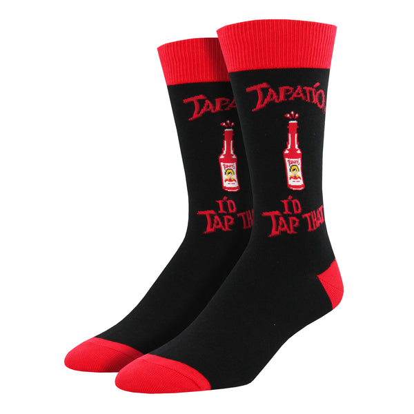Shown on a foot form, a pair of Socksmith's black cotton men's crew socks with red cuff/heel/toe, Tapatio hot sauce bottle and the text “Tapatio: I’d Tap That!”