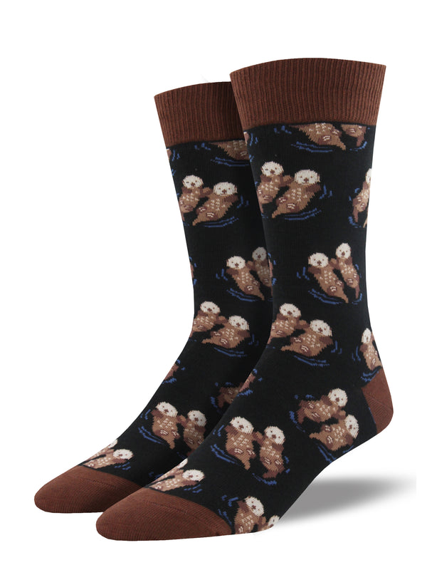 Shown on a leg form, these black cotton men's crew socks with a brown heel, toe and cuff by the brand Socksmith feature adorable otters floating in the ocean holding hands.