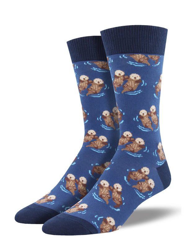 Shown on a leg form, these blue cotton men's crew socks with a navy heel, toe and cuff by the brand Socksmith feature adorable otters floating in the ocean holding hands.