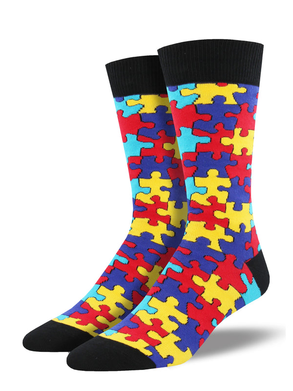 Shown on a foot form, a pair of Socksmith's cotton men's crew socks with black cuff/heel/toe and large all-over pattern of puzzle pieces in blue, red and yellow