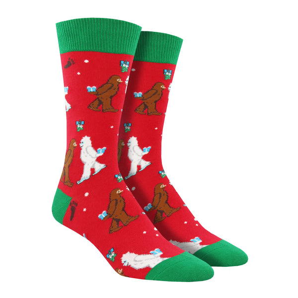 Shown on leg forms, a pair of men's Socksmith brand red cotton crew socks with a green heel, toe, and cuff. These socks feature a brown and white yeti meeting under mistletoe each with little gifts behind their backs.