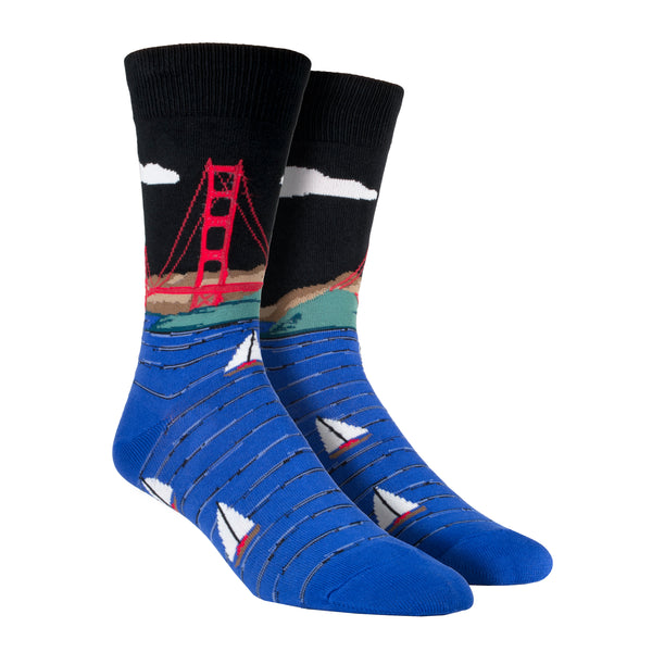 Shown on a leg form, these black cotton men's crew socks by the brand Socksmtih feature the iconic landmark of San Francisco, the Golden Gate Bridge, towering above the beautiful blue San Francisco Bay filled with sailboats.