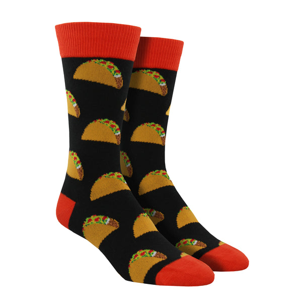 Shown on a foot form, a pair of Socksmith's black cotton men's crew socks with orange cuff/heel/toe and all-over pattern of delicious hardshell tacos