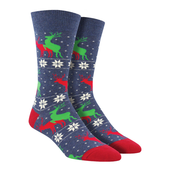 Shown on leg forms, a pair of men's Socksmith brand cotton crew sock in blue with a red heel and toe. This sock features a snowflake sweater pattern stripe with 2 reindeer, one red and one green, mounted and mating.