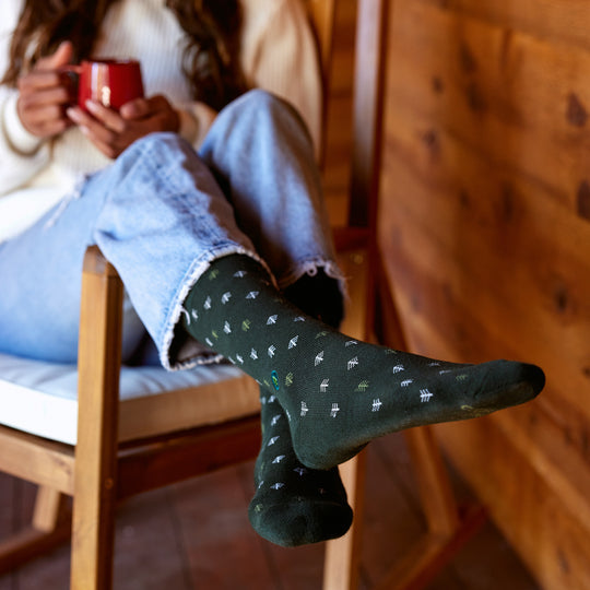 a woman in a wood paneled room holding a mug and wearing jeans and a pair of green conscious step socks with small trees all over them