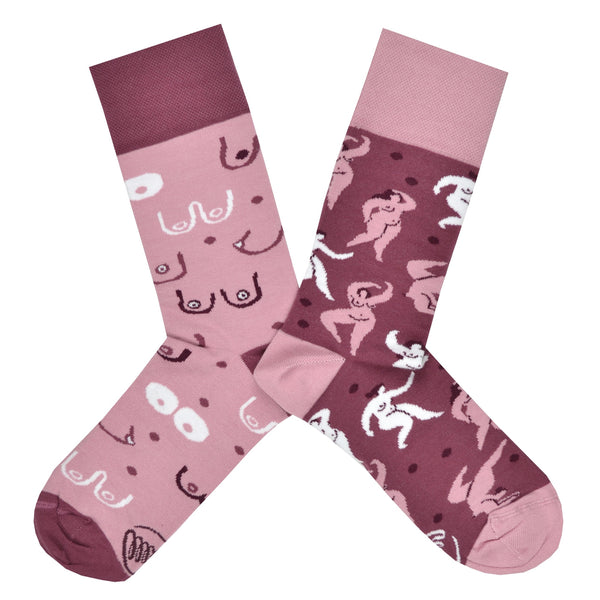 Shown in a flatlay, a pair of mismatched Many Mornings brand unisex cotton crew socks. One sock has a dark pink heel/cuff/toe with a light pink base and cartoon images of different breast shapes. The second sock has light pink heel/cuff/toe and a darker pink base features an all over design of curvaceous nude feminine bodies.