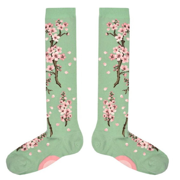 Shown in a flatlay, a pair of Mod Socks brand women's cotton knee high socks in light green. The sock features cherry blossom branches going from the foot to the top of the sock with pink flowers and loose pink petals dot the rest of the sock.