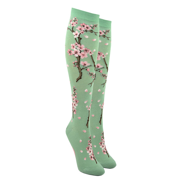 Shown on leg forms, a pair of Mod Socks brand women's cotton knee high socks in light green. The sock features cherry blossom branches going from the foot to the top of the sock with pink flowers and loose pink petals dot the rest of the sock.