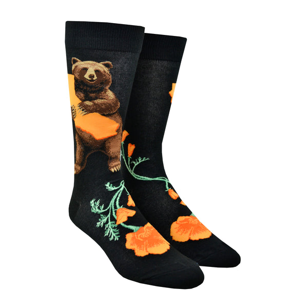 Shown on a leg form, these black cotton men's crew socks by the brand Mod Socks feature a bear standing and hugging a large orange sketch of the state of California, and orange California poppy's on the foot.