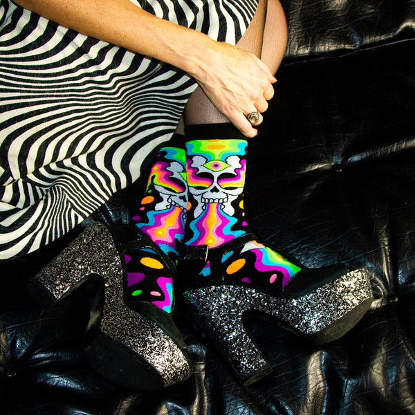 A model wearing a psychedelic black and white dress, sparkly chunky heels, and a pair of Gumball Poodle black cotton crew socks with illustrative skull spewing neon rainbow psychedelic spirals all down the foot