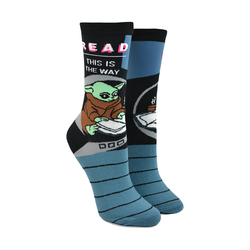 Shown on a leg form, these blue and black cotton unisex crew socks by the brand Out of Print feature the Star Wars Mandalorian character Baby Yoda reading, with the text 