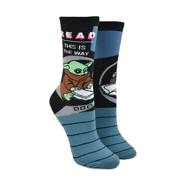 Shown on a leg form, these blue and black cotton unisex crew socks by the brand Out of Print feature the Star Wars Mandalorian character Baby Yoda reading, with the text "Read This Is The Way" written on the top of the leg.