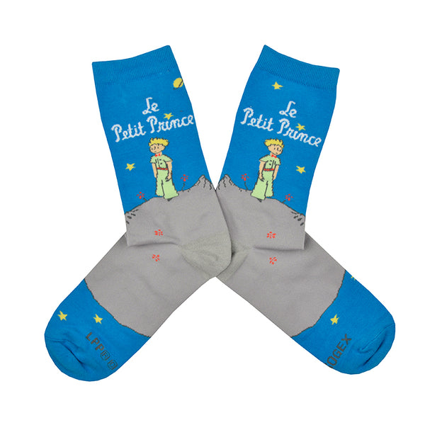Shown in a flatlay, a pair of Out of Print brand unisex cotton crew socks in blue and grey. These socks feature a little cartoon boy outside at night with the words, "Le Petit Prince" on the leg. The image is take from the 1943 classic children's novella The Little Prince written and illustrated by Antonie de Saint-Exupery.