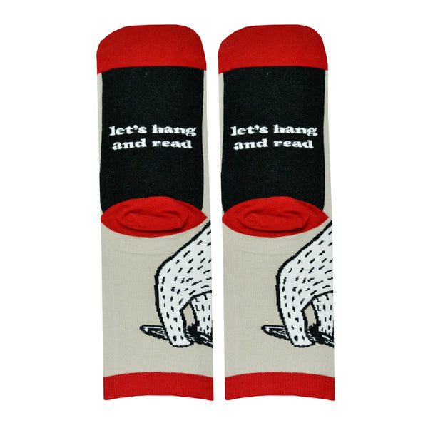 Shown in a flatlay with the sole of the sock faced, a pair of Out of Print brand unisex cotton crew socks in tan with a red heel/toe/cuff and a black sole which reads, "let's hang and read". The sock features a black and white cartoon sloth hanging from a vine with a red book.