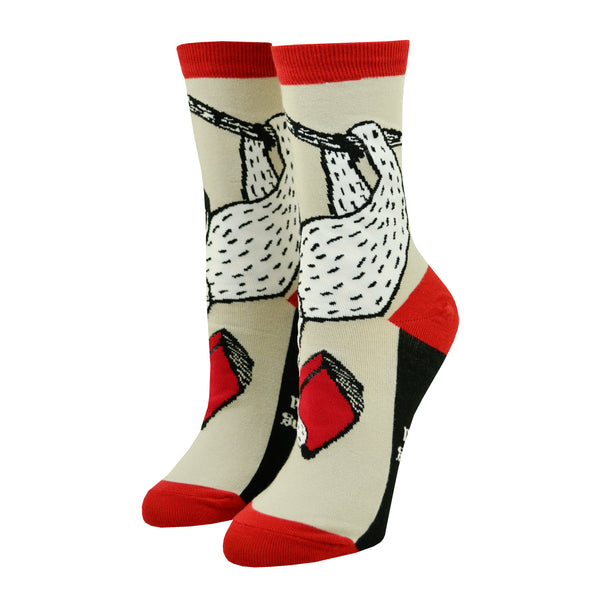 Shown in the smaller size on leg forms from the right, a pair of Out of Print brand unisex cotton crew socks in tan with a red heel/toe/cuff and a black sole. The sock features a black and white cartoon sloth hanging from a vine with a red book.