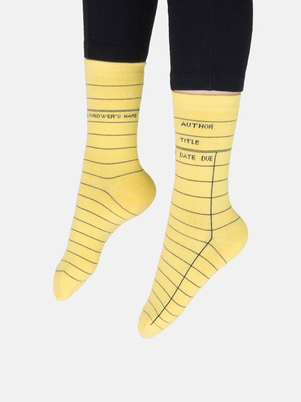 A model wearing yellow cotton unisex crew socks by the brand Out of Print feature the iconic library card design with the words "Author, Title, Date Due" written on the leg and a grid for filling in the information running down the leg and foot.