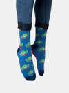 Shown close up on a model, a pair of unisex Out of Print blue cotton crew socks with green goofy faces sticking out their tongue like on the book cover, “Don’t” on the sole of one foot, “Panic” on the other, and navy cuff/heel/toe