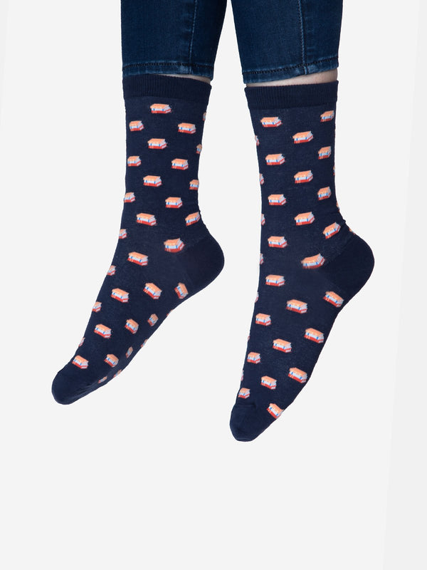 Shown on a models feet, a pair of unisex Out of Print brand cotton crew sock in navy blue with an all over motif of little stacks of books.