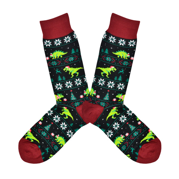 Shown in a flatlay, a pair of men's Socksmith brand cotton crew socks in black with a brick red heel, toe, and cuff. This sock has a Christmas sweater-like design with snowflakes and hat and scarf clad green dinosaurs.