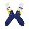 Shown in a flatlay, a pair of Socksmith's cotton men's crew socks with Corona beer logo and signature navy blue and white background