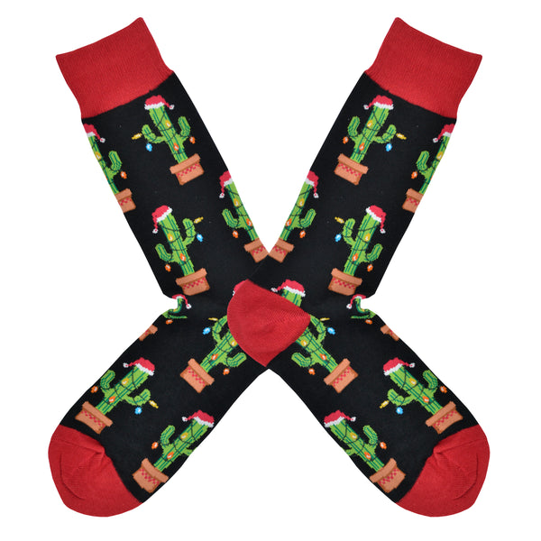 Shown in a flatlay, a pair of men's Socksmith brand cotton crew sock in black with a red heel, toe, and cuff. The sock features an all over motif of potted cactus plants with a string of lights and a Santa hat.