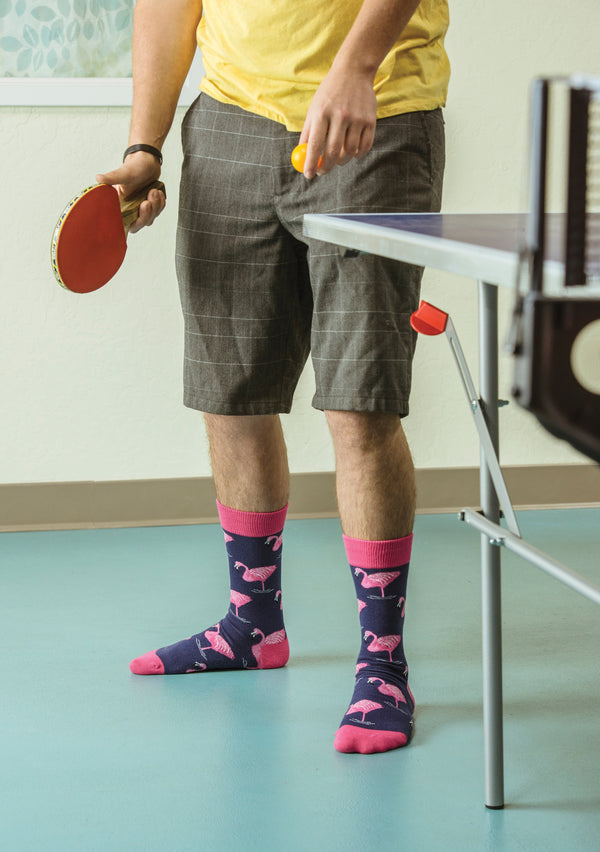 Shown on a model, a pair of Socksmith's navy cotton men's crew socks with pink flamingo all over pattern