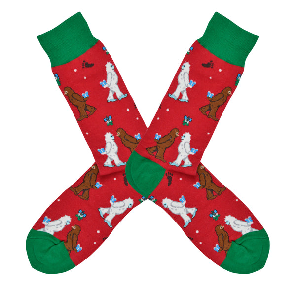 Shown in a flatlay, a pair of men's Socksmith brand red cotton crew socks with a green heel, toe, and cuff. These socks feature a brown and white yeti meeting under mistletoe each with little gifts behind their backs.