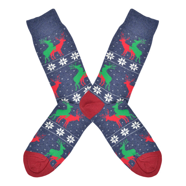Shown in a flatlay, a pair of men's Socksmith brand cotton crew sock in blue with a red heel and toe. This sock features a snowflake sweater pattern stripe with 2 reindeer, one red and one green, mounted and mating.