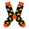 Shown in a flatlay, a pair of Socksmith's black cotton men's crew socks with orange cuff/heel/toe and all-over pattern of delicious hardshell tacos