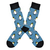 Shown in a flatlay, a pair of Socksmith's blue cotton men's crew socks with black cuff/heel/toe and all-over pattern regal emperor penguins