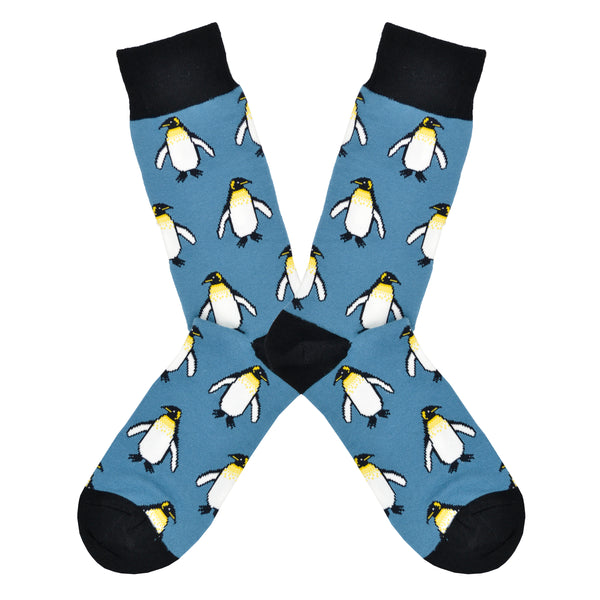 Shown in a flatlay, a pair of Socksmith's blue cotton men's crew socks with black cuff/heel/toe and all-over pattern regal emperor penguins
