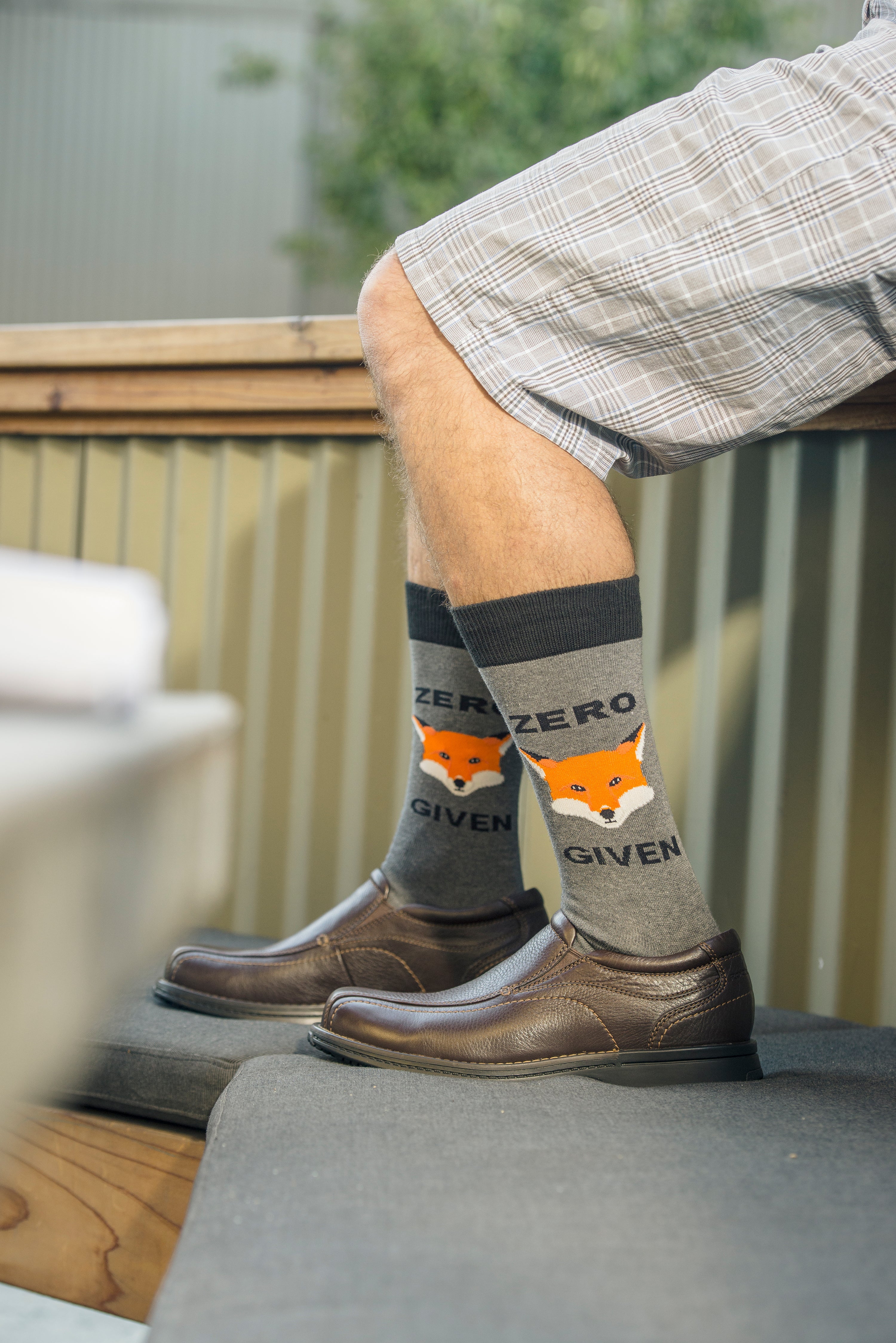 Shown on a light skinned masculine models legs with shoes , a pair of Socksmith brand men's cotton crew socks in grey with black heel/cuff/toe and a cartoon fox face on the leg. The text on the sock reads, 