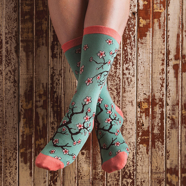 Shown on a light-skinned model against a roughed up woden floor, a pair of Socksmith brand bamboo women's crew sock with a cherry blossom design.
