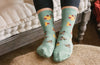 Shown on a model, a pair of Socksmith’s light green cotton women’s crew socks with bee print including yellow honeycombs