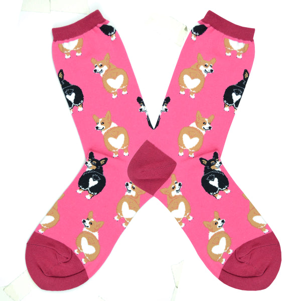 Shown in a flatlay, a pair of Sock Smith brand women's cotton crew socks in hot pink with a magenta heel, toe, and cuff. The sock features an all over motif of blonde and black corgi dogs with their little heart shaped butts facing out.