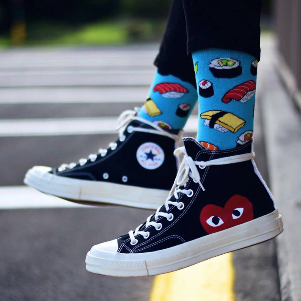 A model wearing Converse and a pair of Socksmith's sky blue cotton men's crew socks with various types of sushi rolls and chopsticks in an all-over pattern