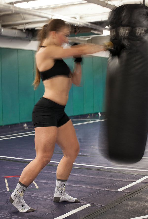 A strong looking woman in active wear is pictured in this blurry image punching a punching bag. On her feet are the grey "Fuck this Shit" socks. 
