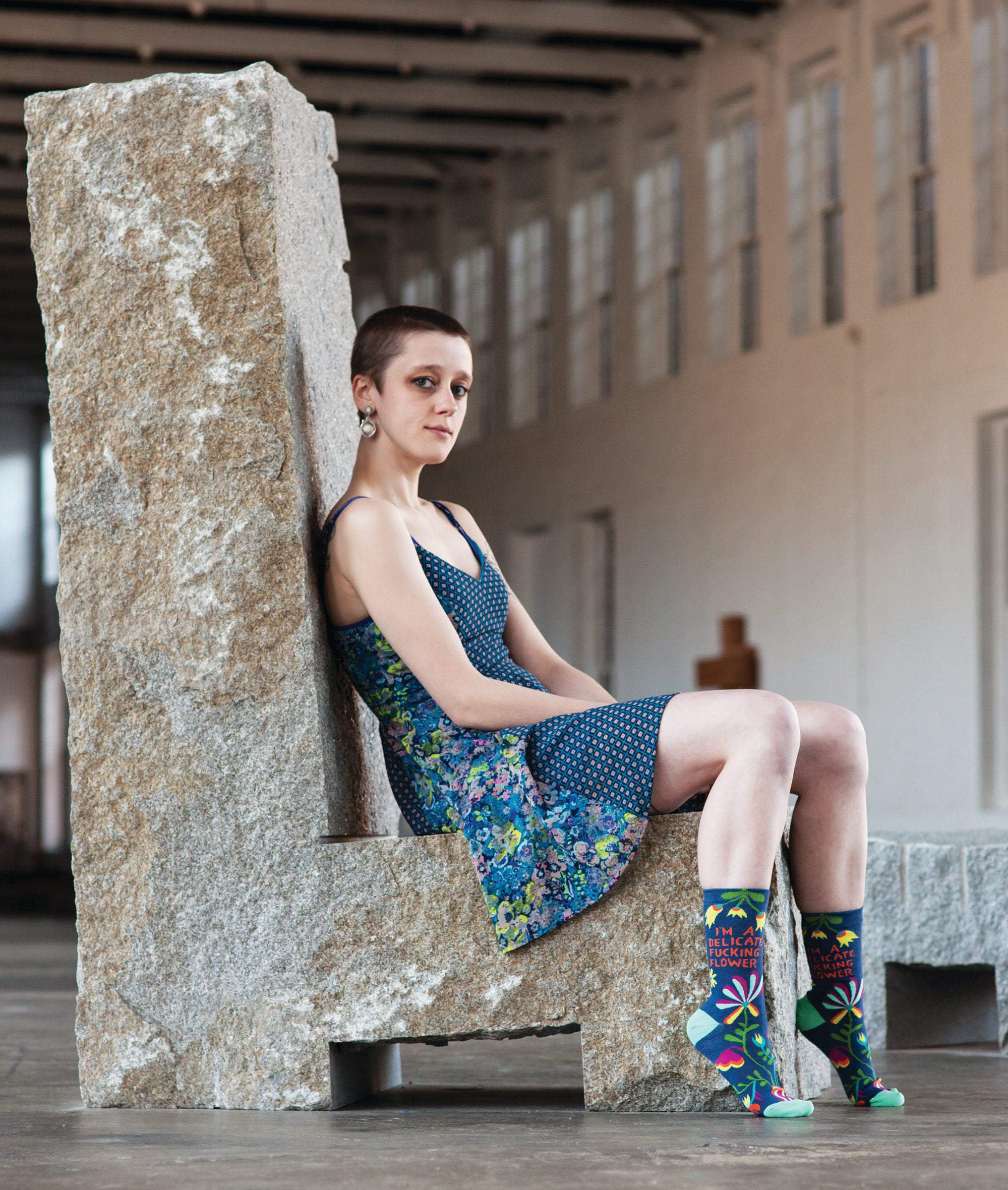 A short haired model in a blue floral dress sits on a cement chair while wearing the 
