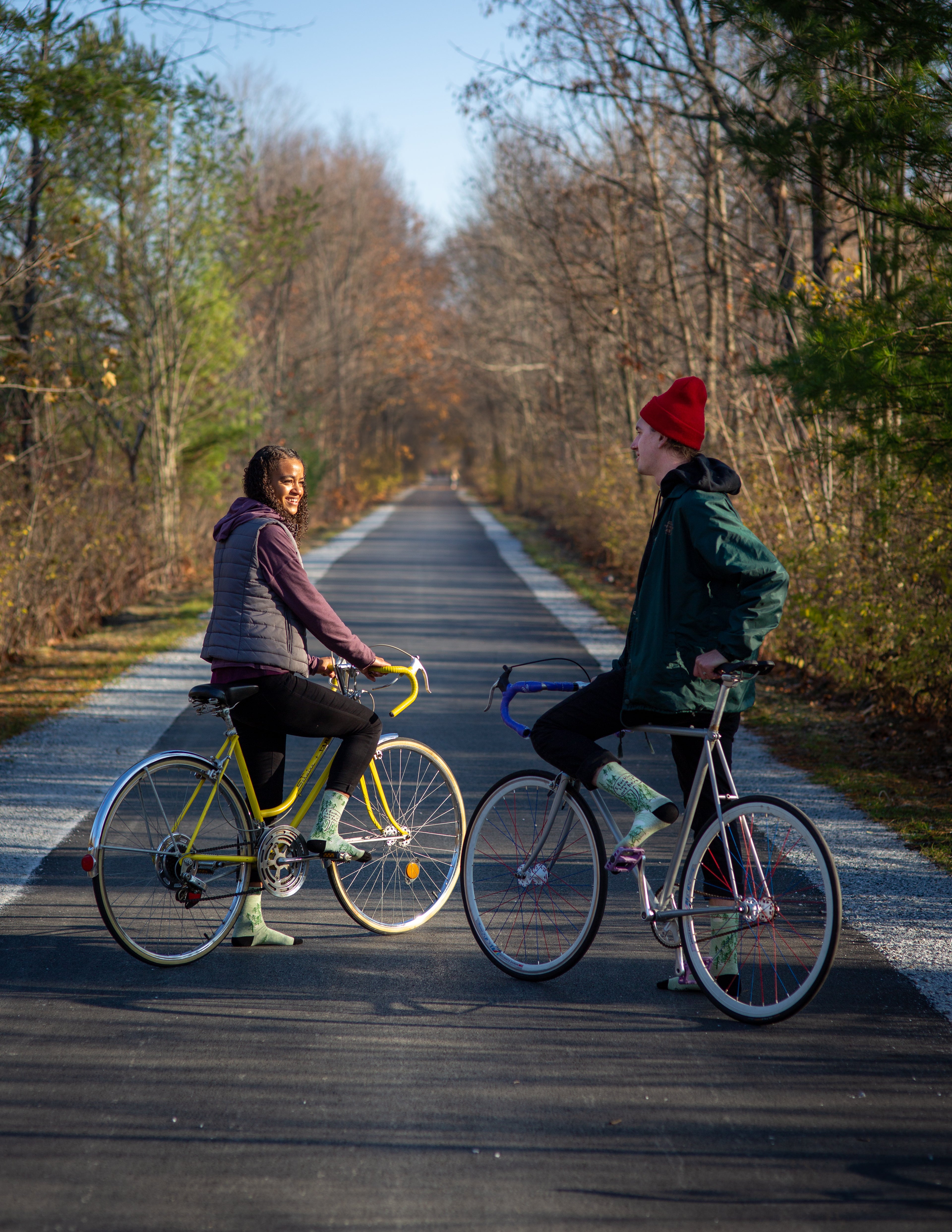 Two people on their bikes in the forest smile at each other. One model is a dark skinned women and the other is a white man. They are both wearing the 