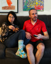 an interracial couple sits on a comfy couch while the man watches TV and the woman has a mischievous look on her face next to him. She is wearing the, " I Never Fart" socks. 