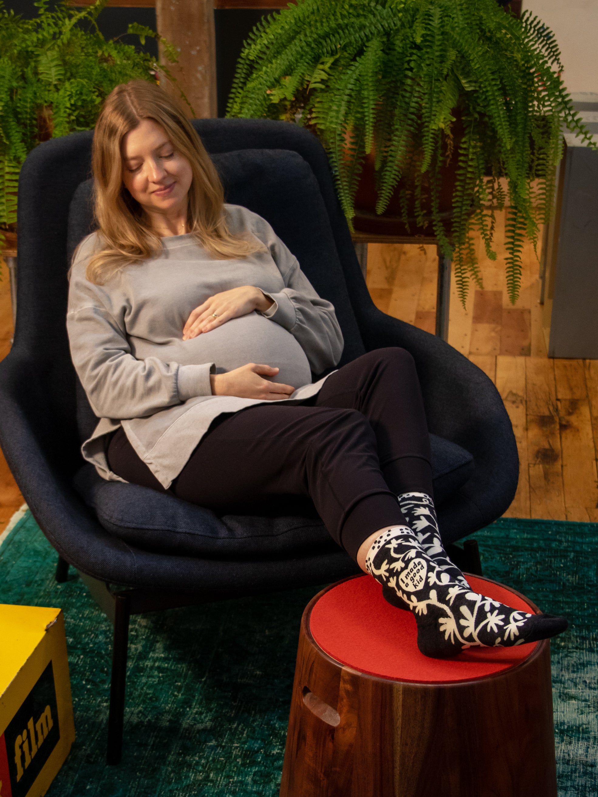 A light skinned pregnant woman lounges on a comfy chair with her feet up. She is cradling her stomach lovingly and wearing the Blue Q 