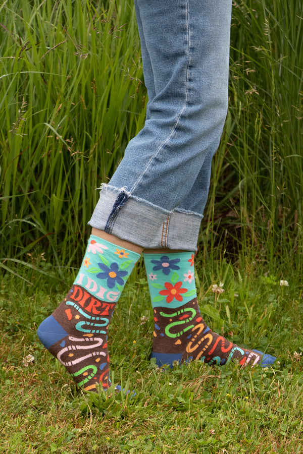 a models legs in jeans and multicolored socks that read i dig dirt featuring colorful flowers and worms, standing on a grassy lawn