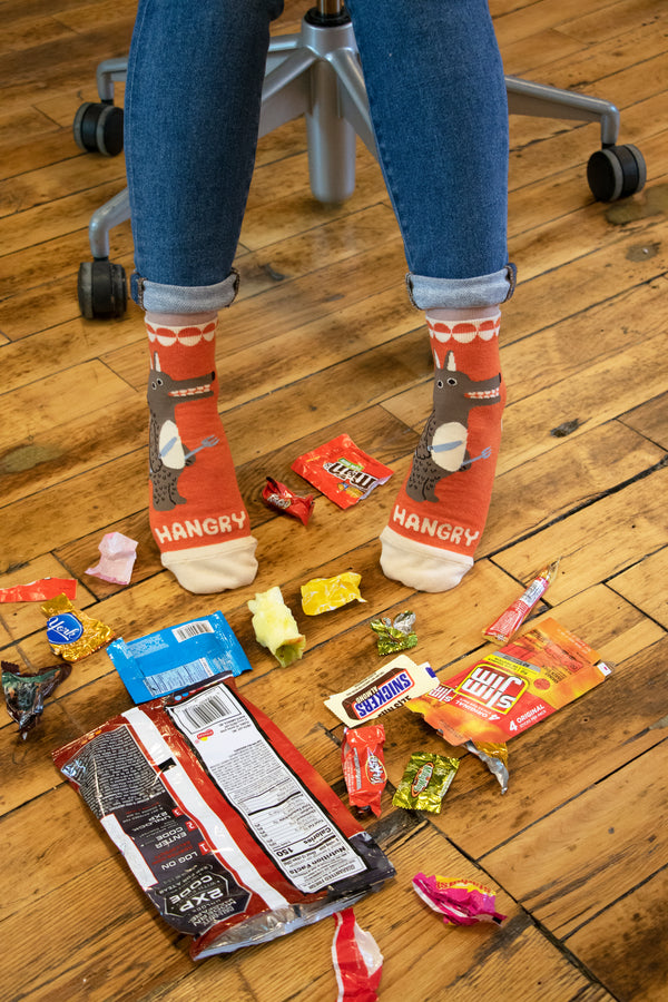 Shown on a models feet, the orange "HANGRY" socks witha bile of snack food wrappers on the floor. 