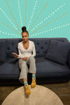 A dark skinned woman is shown on her couch wearing Blue Q psychic socks while holding her phone. On the wall behind her is a dot design giving the illusion that she is seeing more than we know. 