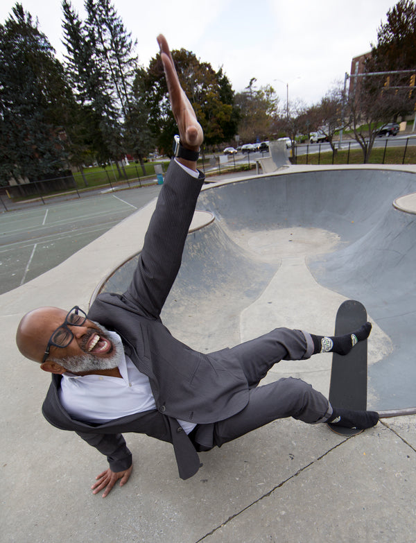 An older African American man is shown doing a skateboard trick in the Blue Q socks. He is wearing a suit and  has an excited look on his face.